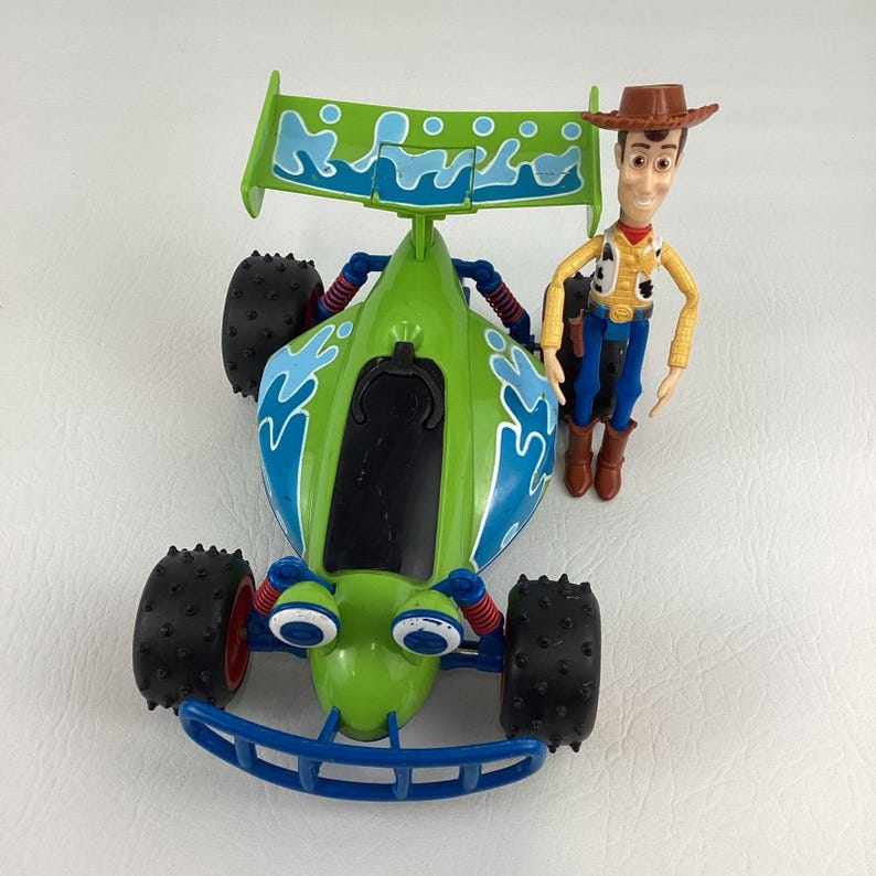 May include: A green and blue toy race car with large black tires and a Woody action figure. The car has a spoiler with a wave design. Woody is wearing a cowboy hat, vest, and blue jeans.
