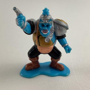 May include: A blue and silver action figure of a creature with a large head, holding a gun. The figure is standing on a blue base.