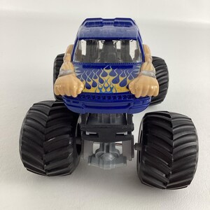 May include: A blue toy monster truck with large black tires. The truck has a flame design in yellow and blue on the front. The truck has a cartoonish design with arms and hands.