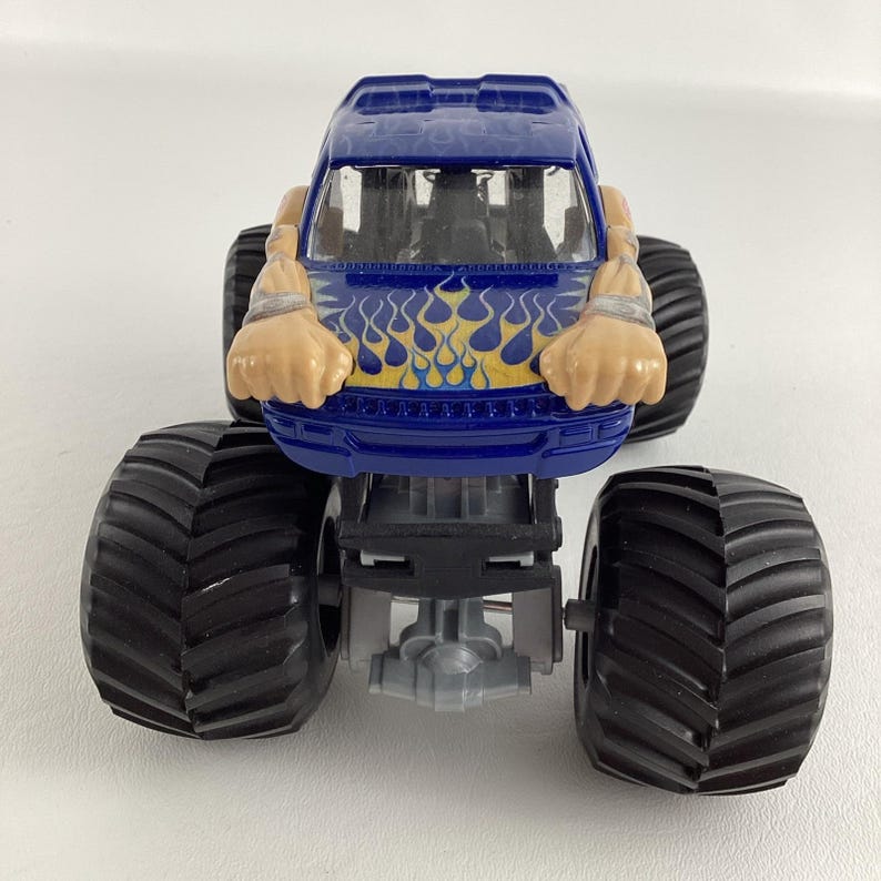 May include: A blue toy monster truck with large black tires. The truck has a flame design in yellow and blue on the front. The truck has a cartoonish design with arms and hands.