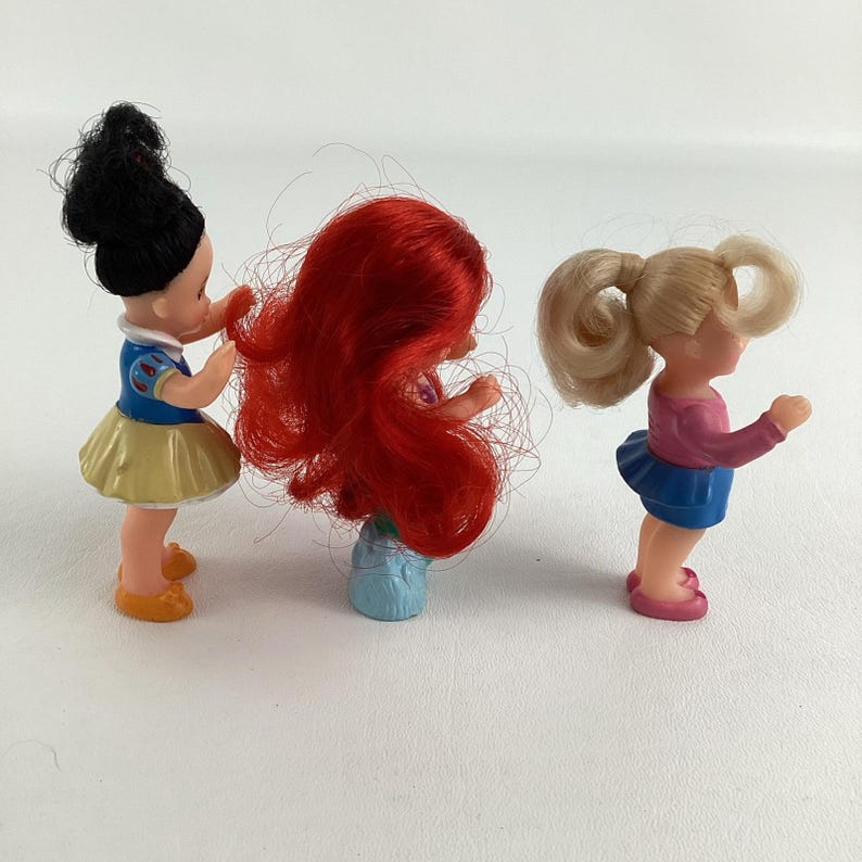 May include: Three small plastic dolls, each with a unique hairstyle and outfit. One doll has black hair and a yellow skirt, another has red hair, and the third has blonde hair and a blue skirt. The dolls are posed as if interacting.
