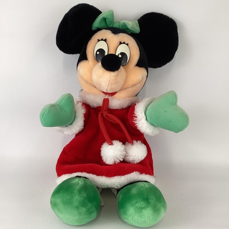 Puede incluir: Un peluche de un personaje de rat&oacute;n con un vestido de terciopelo rojo con ribete de piel sint&eacute;tica blanca, manoplas verdes y zapatos verdes. El personaje tiene orejas negras grandes, un lazo verde claro y un cord&oacute;n rojo con dos pompones.
