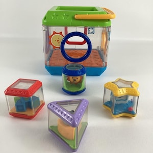 May include: A clear plastic cube with a handle and a variety of colorful shapes inside. The cube has a blue, green, and yellow design. The shapes include a star, a triangle, a square, and a cylinder. The cylinder has a lion face on it.