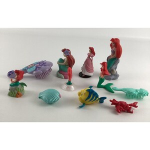 May include: A collection of colorful figurines featuring characters from a popular animated film. The set includes mermaid figures with red hair, a yellow fish, a red crab, and other themed accessories. The figurines are made of plastic.