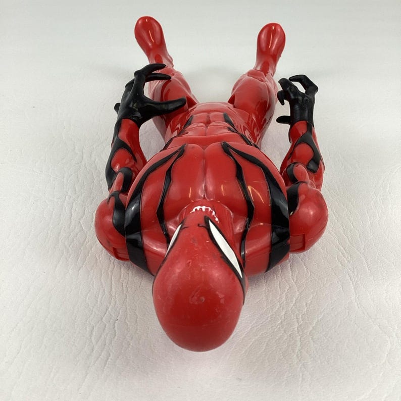 Puede incluir: Una figura de acci&oacute;n roja de un superh&eacute;roe con detalles negros. La figura est&aacute; tumbada boca arriba, con los brazos y las piernas extendidos. La figura tiene una m&aacute;scara blanca y manos negras.