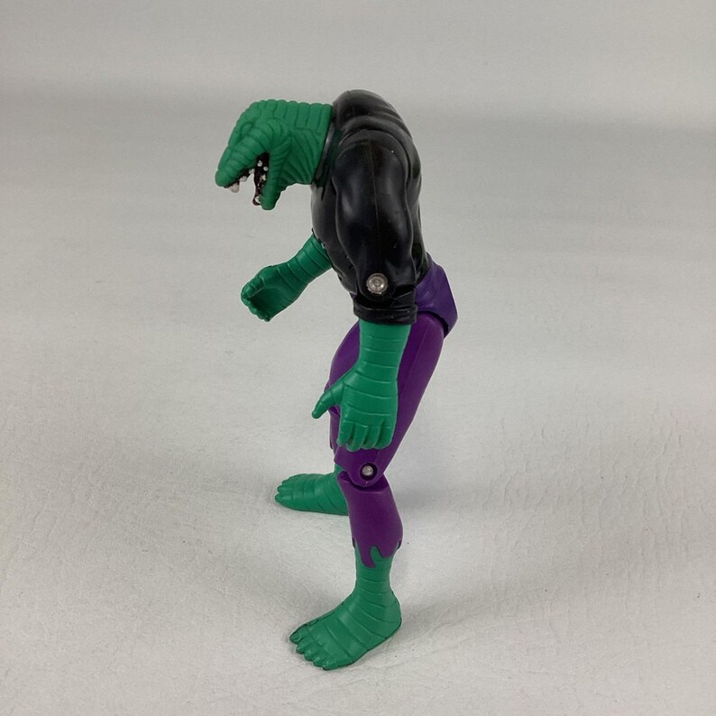 Puede incluir: Una figura de acci&oacute;n con forma de lagarto, con cabeza de reptil texturizada, torso negro y pantalones morados. La figura tiene brazos y piernas verdes con articulaciones visibles en hombros, codos y rodillas, en una postura encorvada.