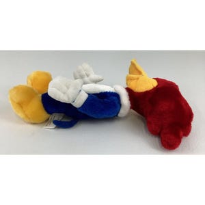 Op de afbeelding: Een pluche speelgoed met een stripfiguur in de kleuren geel, wit, blauw en rood. Het speelgoed heeft witte handschoenen, een blauw lichaam en een rode hand. Het hoofd en de voeten van het personage zijn geel.