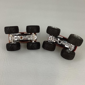 Puede incluir: Dos monster trucks rojos con neum&aacute;ticos negros grandes y chasis plateados se muestran boca abajo. Los neum&aacute;ticos tienen un dibujo profundo. Los camiones est&aacute;n dise&ntilde;ados para el juego todoterreno.