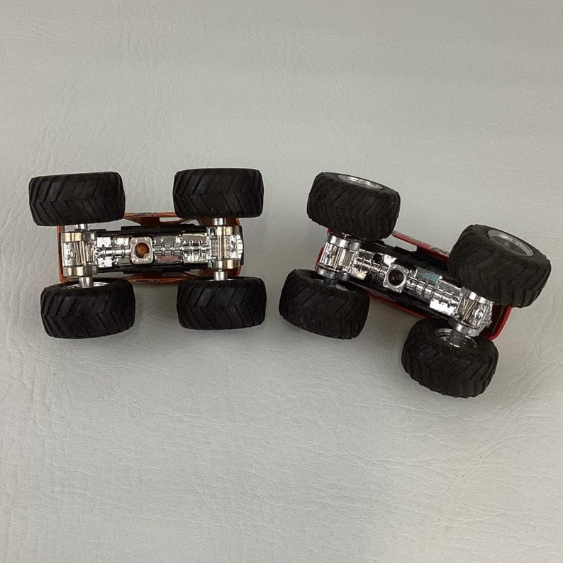 Puede incluir: Dos monster trucks rojos con neum&aacute;ticos negros grandes y chasis plateados se muestran boca abajo. Los neum&aacute;ticos tienen un dibujo profundo. Los camiones est&aacute;n dise&ntilde;ados para el juego todoterreno.