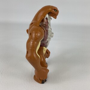 May include: A brown and tan plastic action figure with a reptilian head, muscular arms, and a translucent chest piece. The figure features green eyes and a segmented body design, captured from a side profile view showing its upright posture.