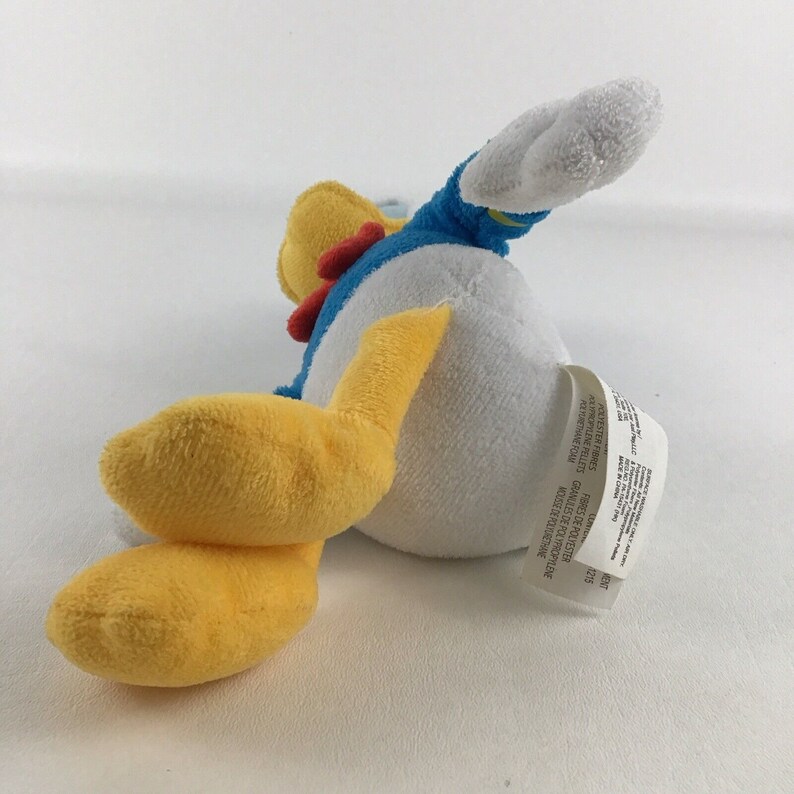 May include: A yellow and white plush toy of Donald Duck wearing a blue sailor shirt with a red bow tie. The toy is lying on its side with its feet in the air.