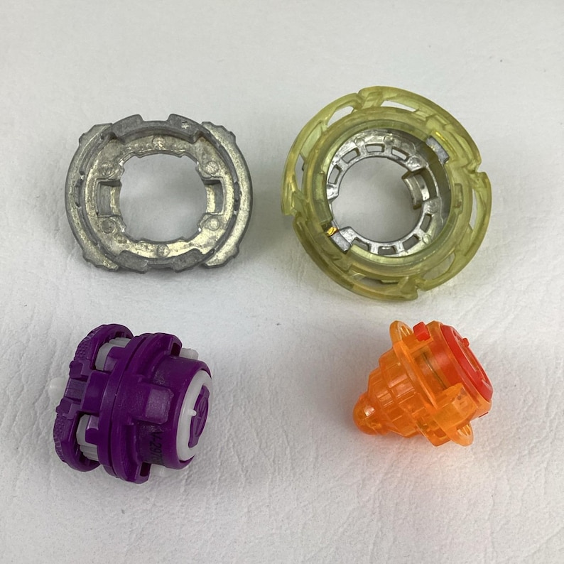 May include: Four disassembled toy components. A silver metal ring, a translucent yellow ring, a purple and white piece, and an orange and red piece. The parts are arranged on a white surface.