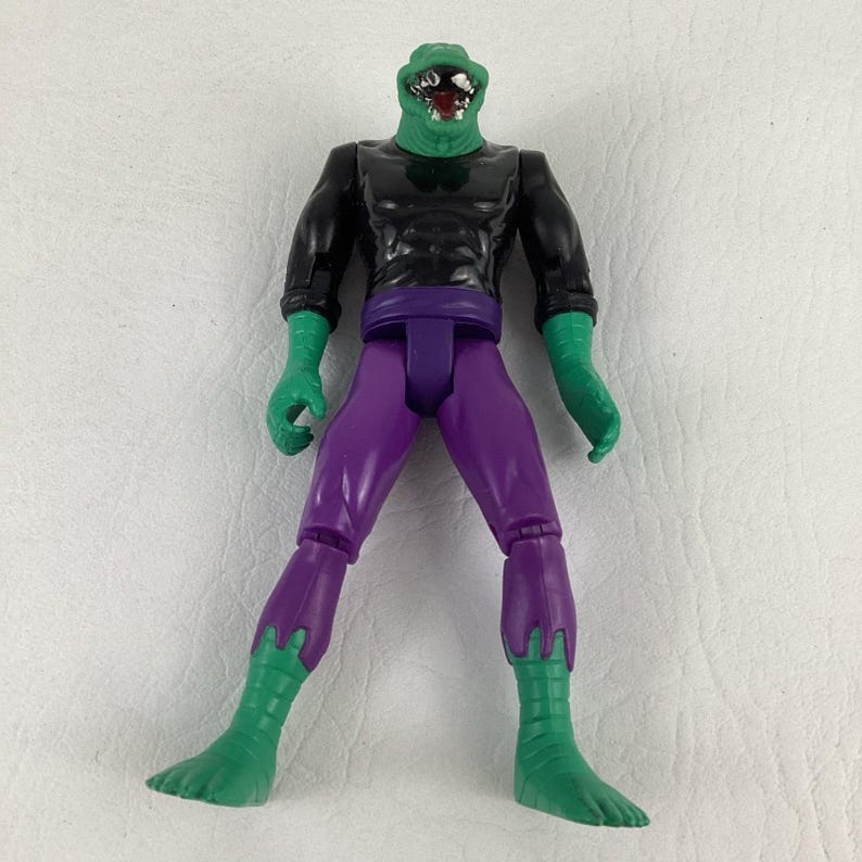 Puede incluir: Una figura de acci&oacute;n de piel verde con cabeza de reptil, dientes afilados y la boca abierta. La figura viste una camiseta negra de manga larga, pantalones morados y guantes y botas verdes. Las extremidades est&aacute;n articuladas en las uniones.