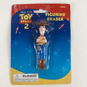 K&ouml;nnte beinhalten: Ein Toy Story 2 Radiergummi in Form einer Woody-Figur in blau-brauner Kleidung, verpackt auf einer blau-roten Karte. Die Verpackung enth&auml;lt das Disney Pixar-Logo und eine Warnung vor Erstickungsgefahr.