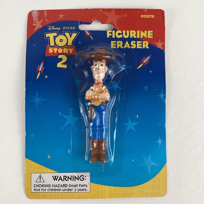 K&ouml;nnte beinhalten: Ein Toy Story 2 Radiergummi in Form einer Woody-Figur in blau-brauner Kleidung, verpackt auf einer blau-roten Karte. Die Verpackung enth&auml;lt das Disney Pixar-Logo und eine Warnung vor Erstickungsgefahr.
