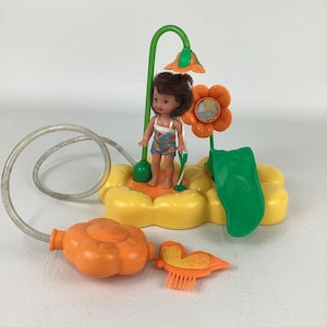 以下が含まれることがあります： 小さな人形が水着姿で黄色い花の形をした台座に立っている、ヴィンテージの人形プレイセット。セットには、緑色のシャワーヘッド、オレンジ色の花、葉、ウォーターホースが含まれています。追加のオレンジ色のアクセサリーも含まれています。