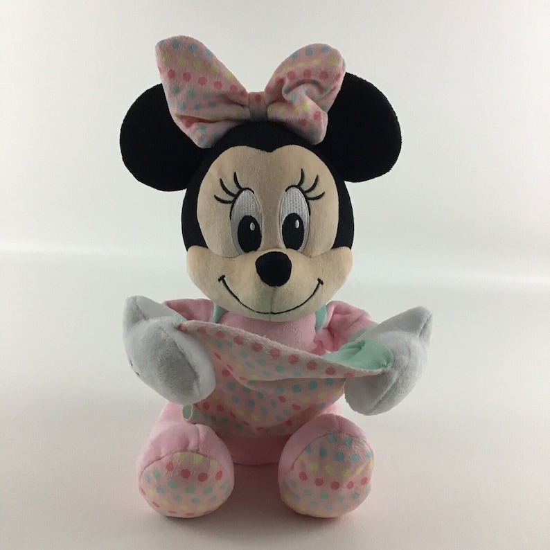 Op de afbeelding: Een roze en wit gestippeld pluche speelgoed van Minnie Mouse met een roze strik. Ze houdt een lichtblauw en groen dekentje vast.