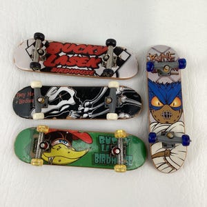 Pode incluir: Quatro mini-skates com vários designs. Um tem o texto "Bucky Laser Birdhouse", outro apresenta um gráfico de esqueleto, um terceiro tem um pato de desenho animado e o último um personagem semelhante a uma múmia. Cada prancha tem rodas pequenas e eixos de metal.