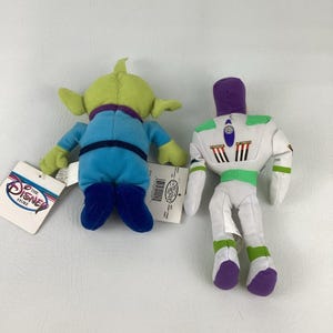 Pu&ograve; includere: Due peluche di The Disney Store. Uno &egrave; un alieno verde a tre occhi con tuta blu e colletto viola. L'altro &egrave; un personaggio ranger spaziale bianco, verde e viola con un design a jetpack sul retro. Entrambi hanno le etichette attaccate.
