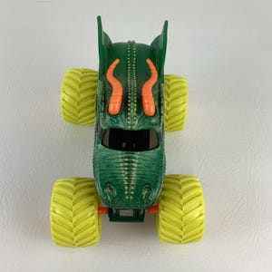 Pu&ograve; includere: Camion mostro giocattolo verde con pneumatici gialli. Il camion presenta un design a forma di drago con accenti arancioni e un parabrezza nero. I pneumatici sono grandi e hanno un motivo a spina di pesce.