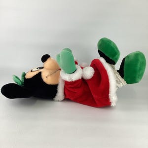 Puede incluir: Un juguete de peluche con cabeza negra y orejas grandes, vestido con un traje rojo con ribete de piel sint&eacute;tica blanca, manoplas verdes y zapatos verdes. El personaje tiene una cara color canela, una nariz negra y un peque&ntilde;o lazo verde.