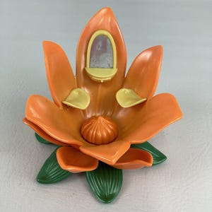 Puede incluir: Juguete de plástico con forma de flor naranja y verde con un pequeño espejo y detalles amarillos. El juguete tiene un diseño detallado y texturizado y es probablemente un artículo decorativo o de juego. La flor tiene hojas verdes en la base.
