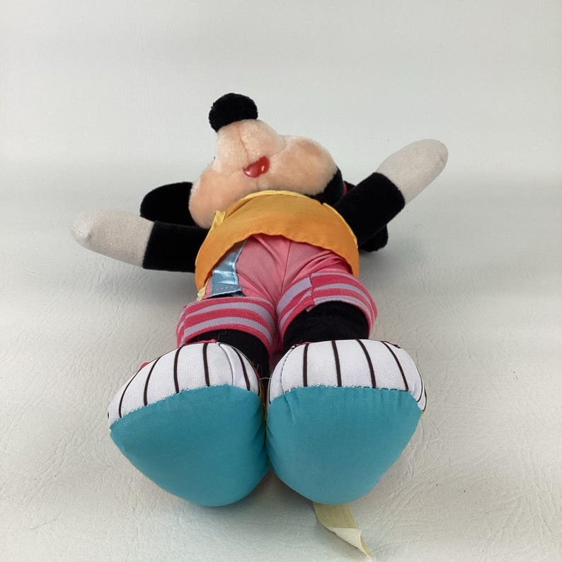 May include: A plush character toy featuring a black head with large round ears, a peach-colored face, and a red tongue. The toy wears a yellow shirt, pink pants with striped cuffs, and white shoes with black vertical stripes and teal soles.