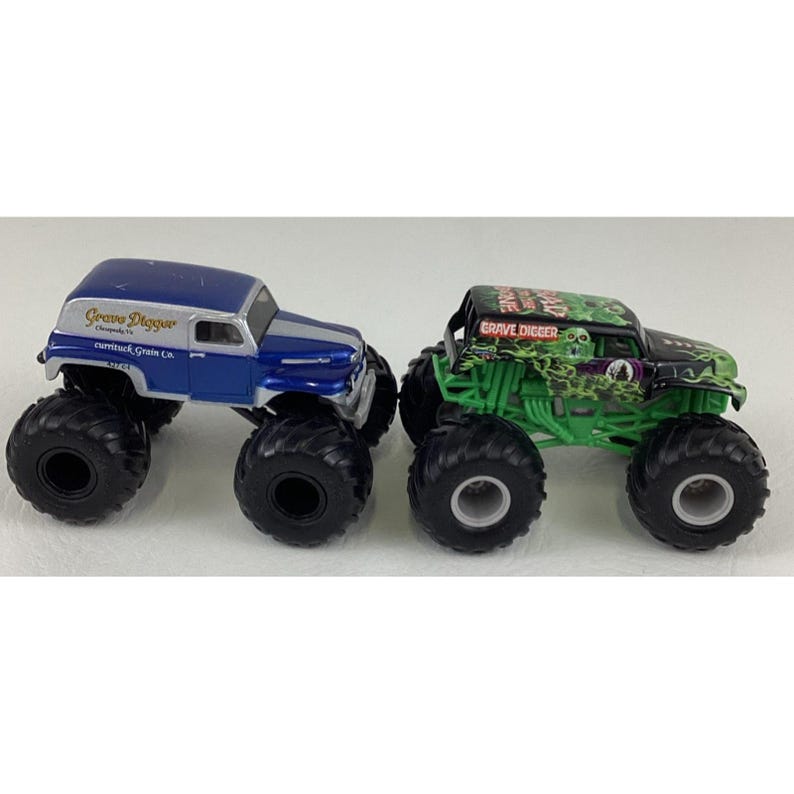 May include: Two toy monster trucks. One is blue and silver with "Grave Digger" text. The other is green and black with "Grave Digger" text and a skull graphic. Both have large black tires.