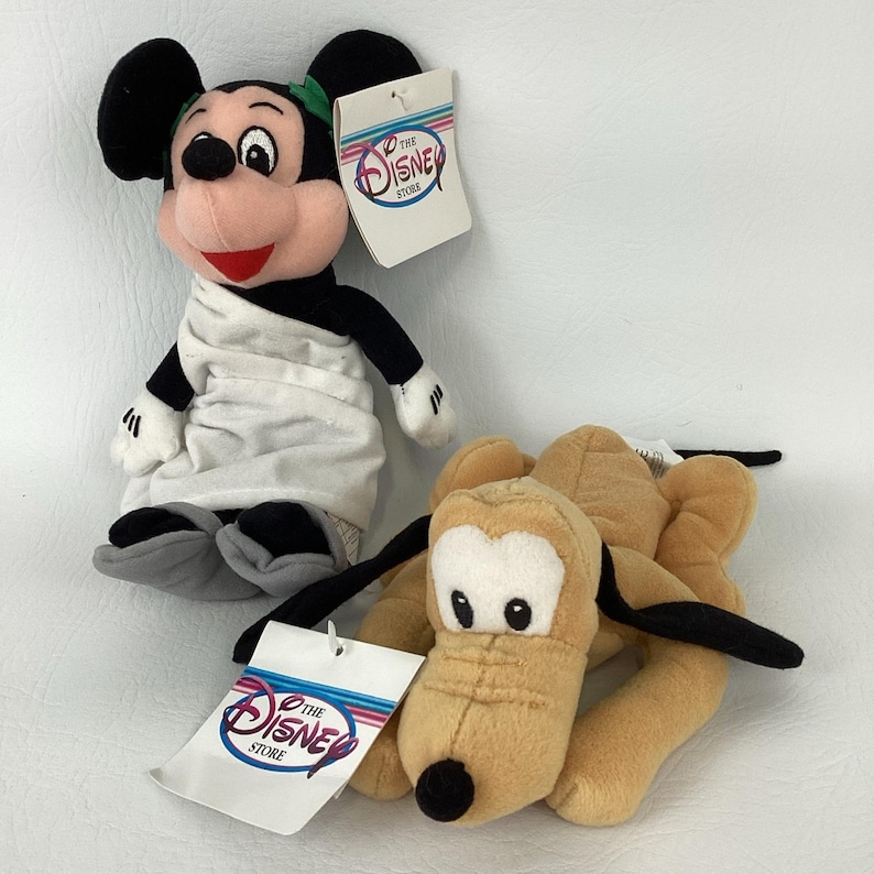 May include: A plush Mickey Mouse doll wearing a white toga and green laurel wreath, accompanied by a plush Pluto dog. Both toys feature The Disney Store tags. Mickey has black ears, a peach face, and grey feet, while Pluto is tan with black ears.
