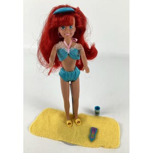 K&ouml;nnte beinhalten: Eine Puppe mit roten Haaren, die einen blauen Badeanzug und ein blaues Stirnband tr&auml;gt. Die Puppe steht auf einem gelben Handtuch mit einem blauen Becher und einem rosa-blauen Kamm.