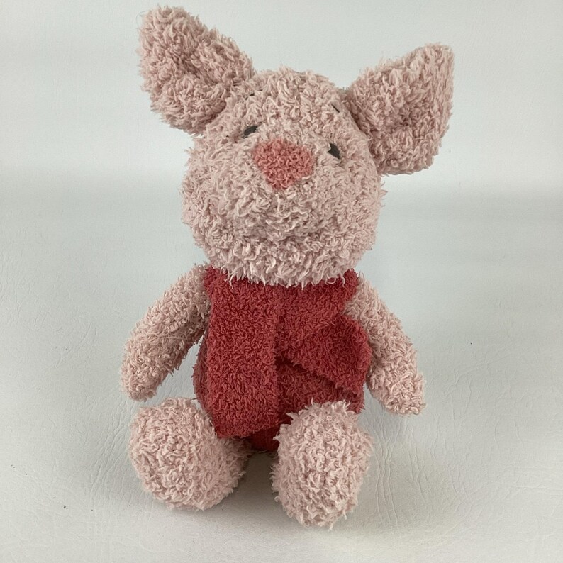May include: A plush toy pig with a pink body, large ears, and a red vest. The pig has a heart-shaped nose and small black eyes. The toy is made of a soft, fuzzy material.