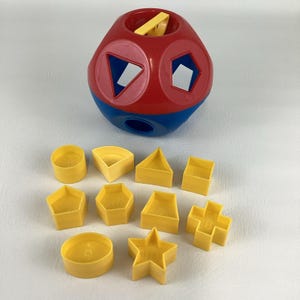 May include: A red and blue shape sorter toy with various yellow geometric shapes. The toy has openings for the shapes, including a circle, triangle, square, and star. The shapes are scattered around the toy.
