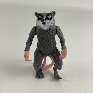 Könnte beinhalten: Grau-schwarze Opossum-Actionfigur mit rosa Schwanz und Händen. Die Figur hat rote Augen und eine lila Nase. Das Opossum steht aufrecht mit den Armen an den Seiten.