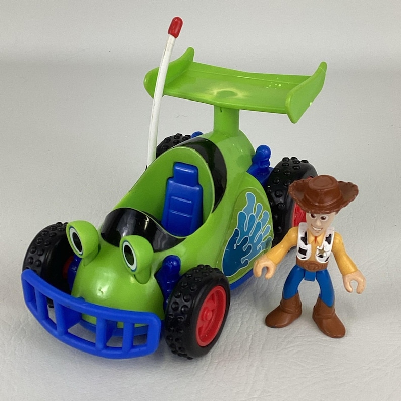 May include: A green and blue toy race car with black tires and red accents, next to a Woody action figure. The car has a spoiler and a remote control antenna. The Woody figure is wearing a cowboy hat and vest.
