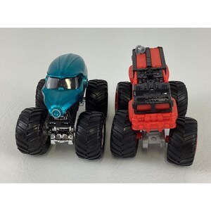 Pu&ograve; includere: Due monster truck giocattolo. Uno &egrave; turchese con un design futuristico, l'altro &egrave; rosso con un design del motore dettagliato. Entrambi hanno grandi pneumatici neri e sono su uno sfondo bianco. Questi camion giocattolo sono progettati per giocare.