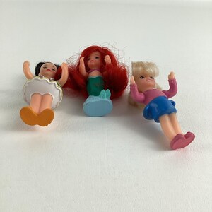 May include: Three small plastic dolls. One doll has black hair and an orange skirt. The second doll has red hair and a green tail. The third doll has blonde hair, a pink shirt, and a blue skirt. All dolls have their arms raised.