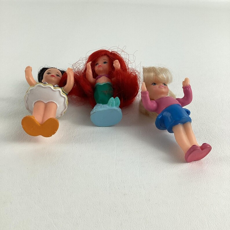 May include: Three small plastic dolls. One doll has black hair and an orange skirt. The second doll has red hair and a green tail. The third doll has blonde hair, a pink shirt, and a blue skirt. All dolls have their arms raised.