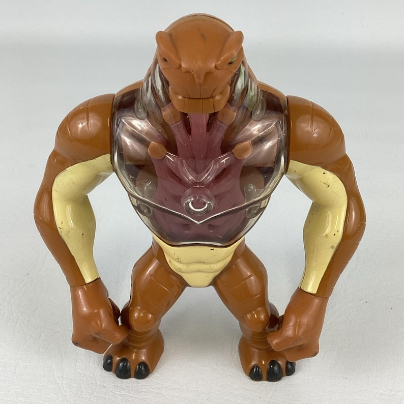 May include: A brown and cream-colored action figure of a creature with a muscular build. The figure features a clear plastic chest plate revealing a dark purple internal structure, a reptilian-like head, and black claws on its feet.