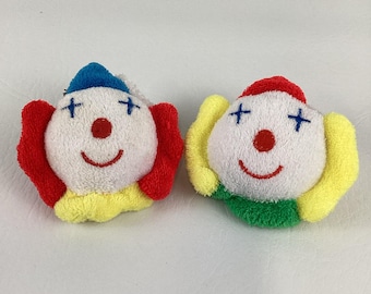 May include: Two plush clown face ornaments. Each has a white face with a red nose and a smiling red mouth. One clown has a blue hat, red ears, and yellow collar. The other has a red hat, yellow ears, and a green collar.