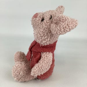 May include: A plush pig toy in shades of pink and red. The pig has a textured, fuzzy appearance, with a long snout and small, dark eyes. It is wearing a red scarf and a matching outfit. The toy is sitting down.