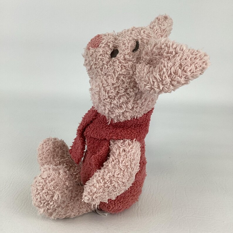 May include: A plush pig toy in shades of pink and red. The pig has a textured, fuzzy appearance, with a long snout and small, dark eyes. It is wearing a red scarf and a matching outfit. The toy is sitting down.