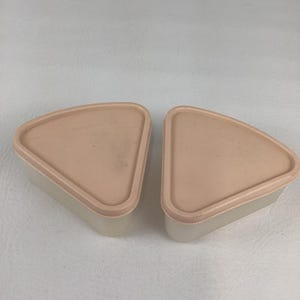 May include: Two vintage Tupperware containers. Each container is a beige triangle shape with a light pink lid. The lids have the Tupperware logo printed on them. The containers are likely for food storage.