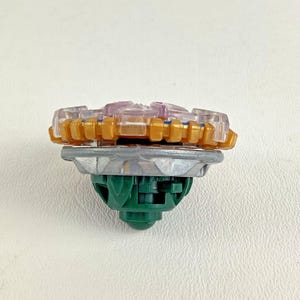 Puede incluir: Un juguete de peonza colorido con una parte superior dorada, transparente y morada, una secci&oacute;n media plateada y una base verde. El juguete est&aacute; dise&ntilde;ado para girar y luchar.