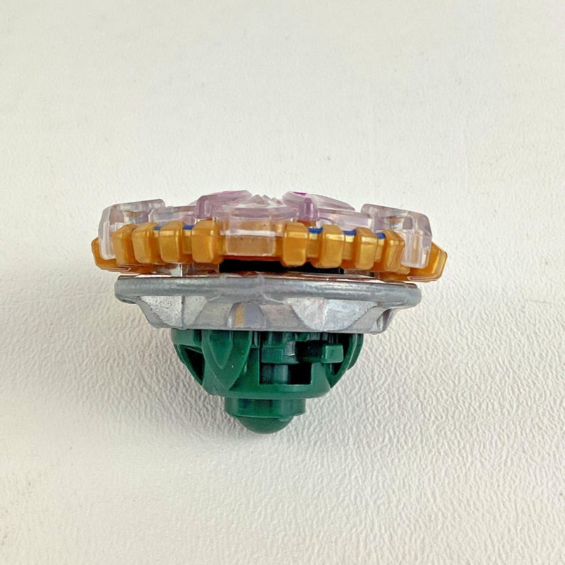 Puede incluir: Un juguete de peonza colorido con una parte superior dorada, transparente y morada, una secci&oacute;n media plateada y una base verde. El juguete est&aacute; dise&ntilde;ado para girar y luchar.