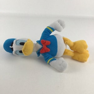 May include: A blue and yellow plush toy of Donald Duck. The toy is lying on its side with its head turned to the left. The toy has a red bow tie and a white belly.