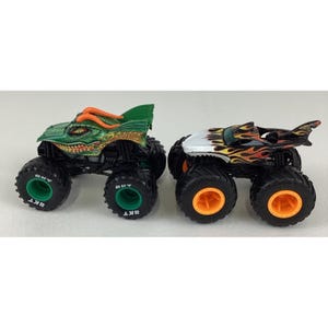 Puede incluir: Dos camiones monstruo de juguete. Uno es verde y naranja, dise&ntilde;ado para parecerse a un drag&oacute;n. El otro es negro, blanco y naranja, con un dise&ntilde;o de aleta de tibur&oacute;n. Ambos tienen neum&aacute;ticos negros grandes con detalles verdes y naranjas.