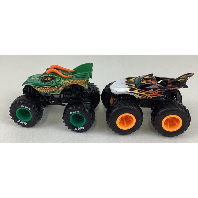 Puede incluir: Dos camiones monstruo de juguete. Uno es verde y naranja, dise&ntilde;ado para parecerse a un drag&oacute;n. El otro es negro, blanco y naranja, con un dise&ntilde;o de aleta de tibur&oacute;n. Ambos tienen neum&aacute;ticos negros grandes con detalles verdes y naranjas.