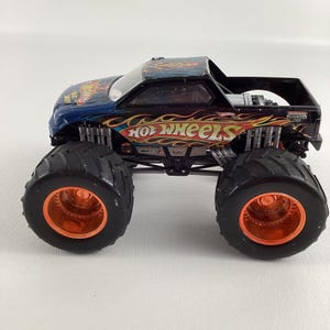 Puede incluir: Un juguete de cami&oacute;n monstruo Hot Wheels negro con ruedas naranjas y negras. El cami&oacute;n tiene un dise&ntilde;o de llama azul y naranja en la cabina y las palabras "Hot Wheels" en naranja. Los neum&aacute;ticos son grandes y negros.