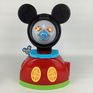 Pu&ograve; includere: Un giocattolo rosso e nero a tema Topolino con un viso rotondo e grandi orecchie nere. Il giocattolo ha un viso blu e bianco con un viso sorridente e due finestre ovali gialle. La base &egrave; rossa con un bordo verde.