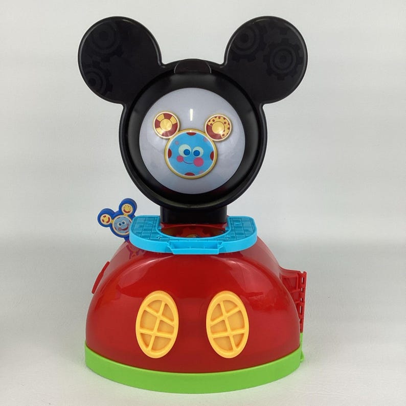 Pu&ograve; includere: Un giocattolo rosso e nero a tema Topolino con un viso rotondo e grandi orecchie nere. Il giocattolo ha un viso blu e bianco con un viso sorridente e due finestre ovali gialle. La base &egrave; rossa con un bordo verde.